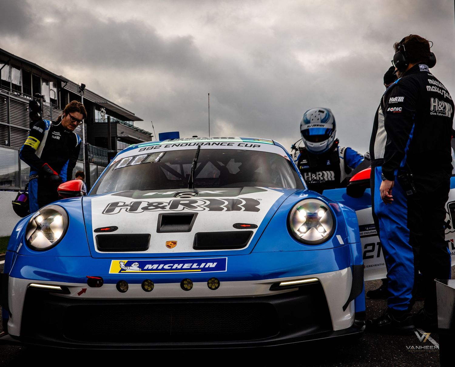 A blue and white Porsche 911 GT3 Cup race car on the grid with mechanics, featuring Michelin sponsorship.