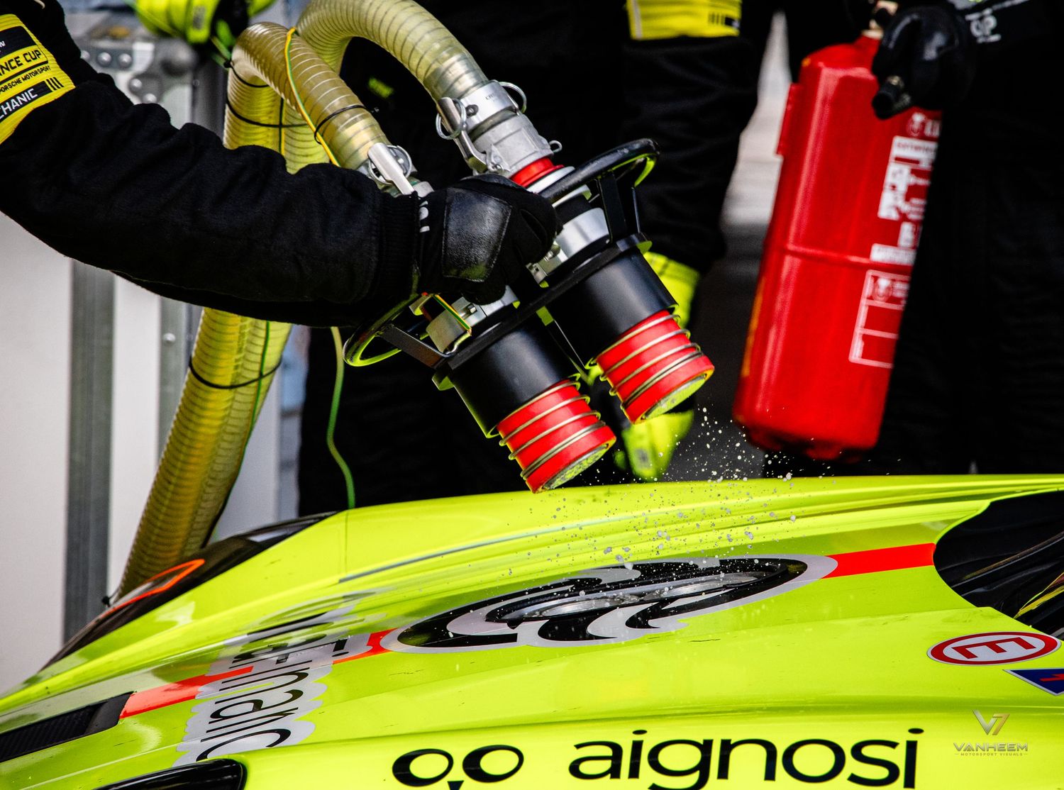 A pit crew member refuels a bright yellow Aignosi racing car during a pit stop, with a red fire extinguisher nearby.