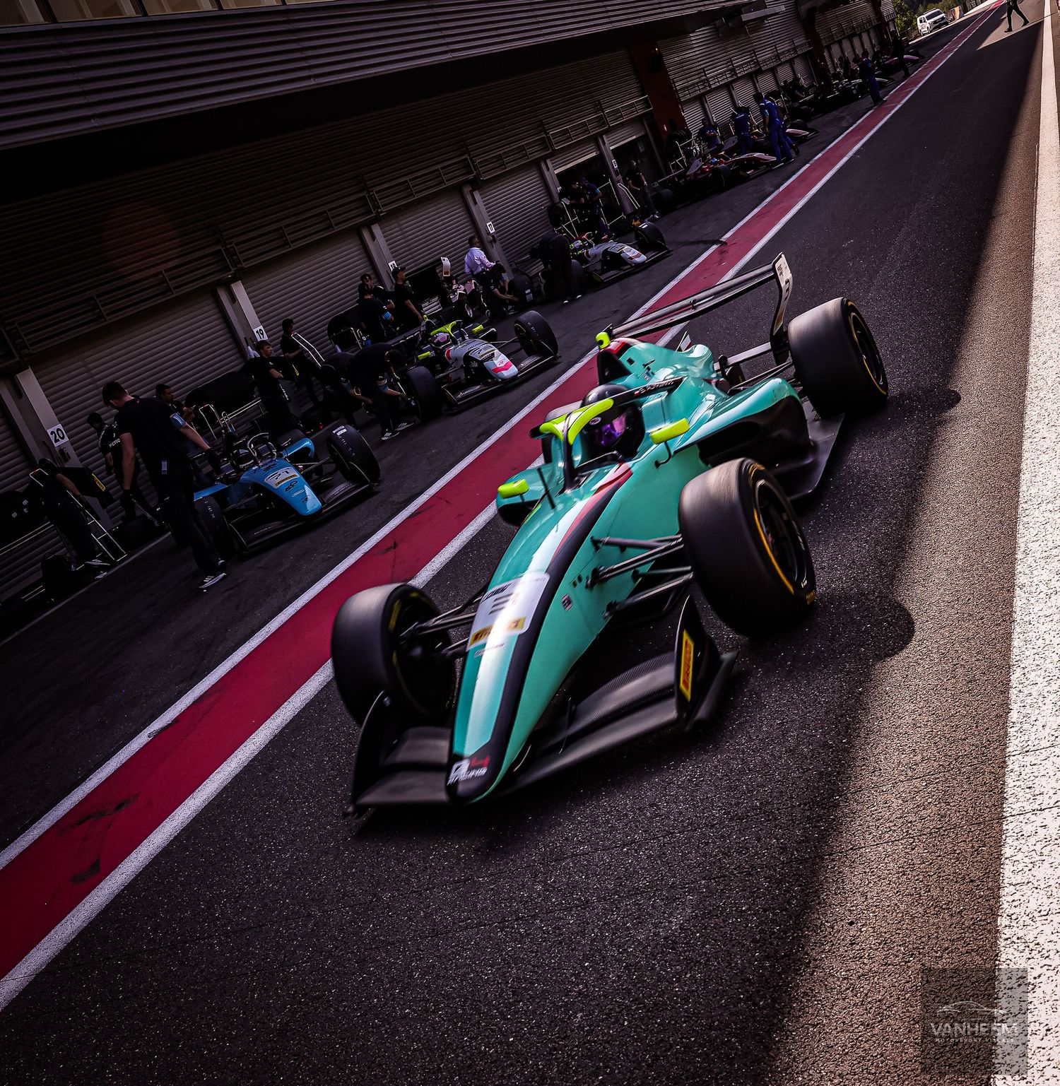 A teal Formula 2 racing car speeds through the pit lane at a circuit, with mechanics and other cars visible in the background.