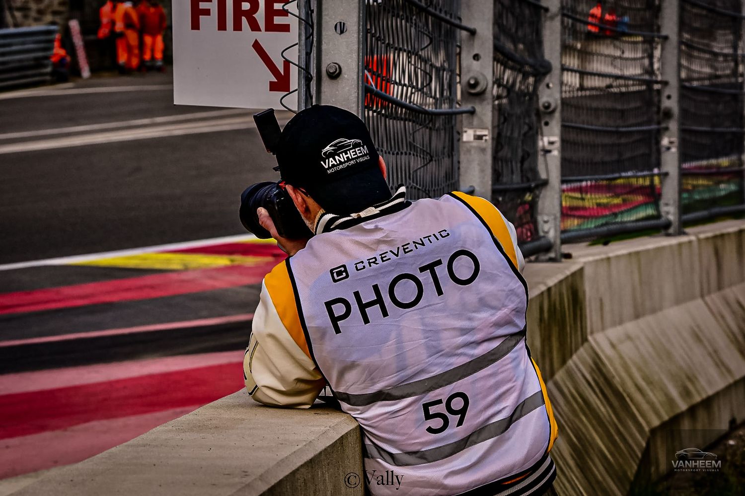 Photographer wearing a yellow PHOTO vest numbered 59 crouches trackside at a Formula 1 race near a FIRE sign.