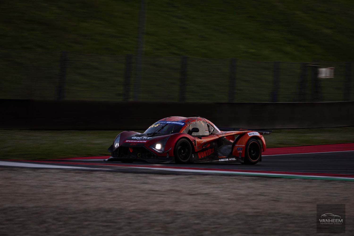 Red race car speeds through a dark racetrack with glowing brake lights during twilight competition.