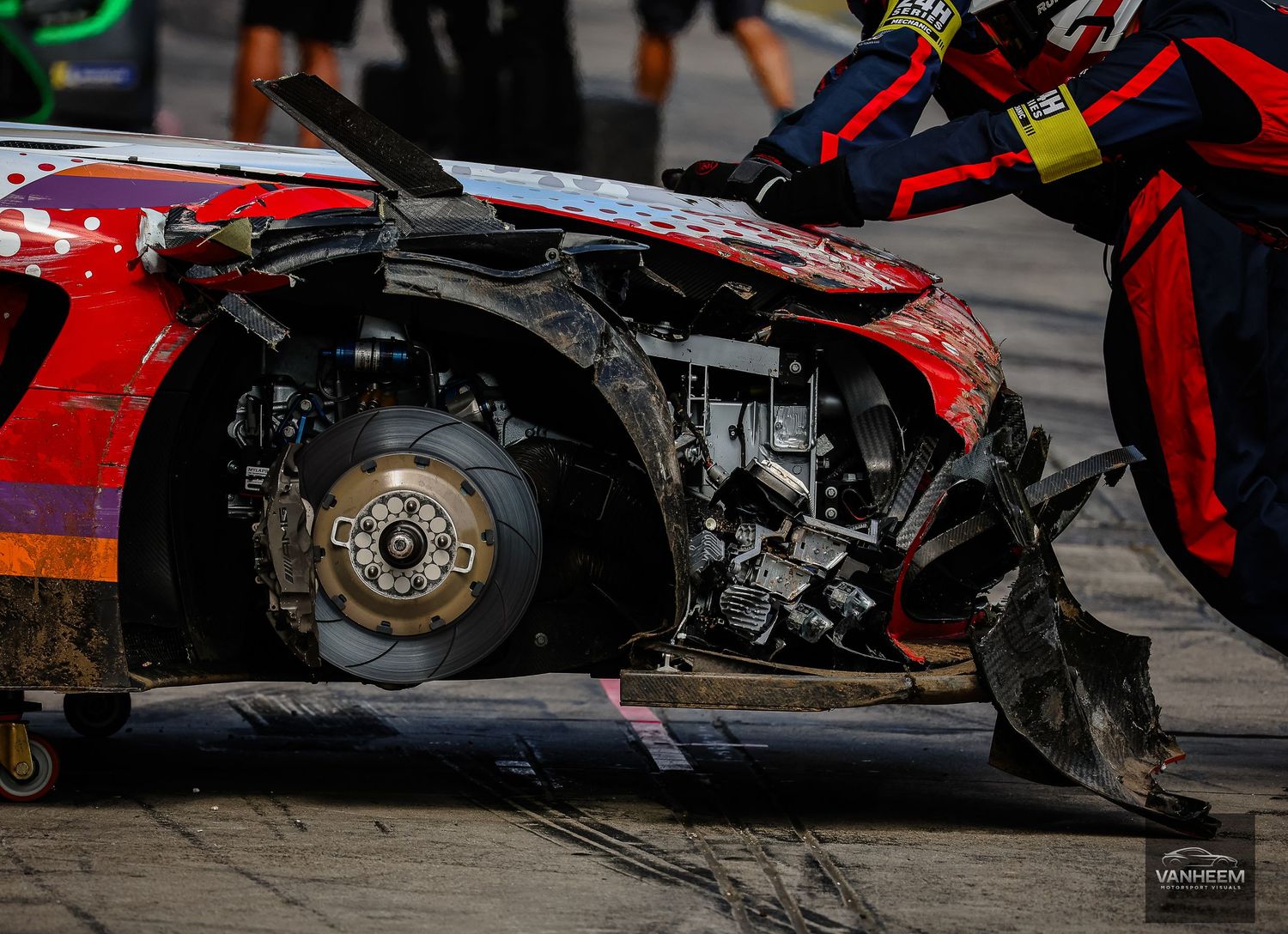 Severely damaged red race car with exposed engine and destroyed front end being recovered on a flatbed at a motorsport event.