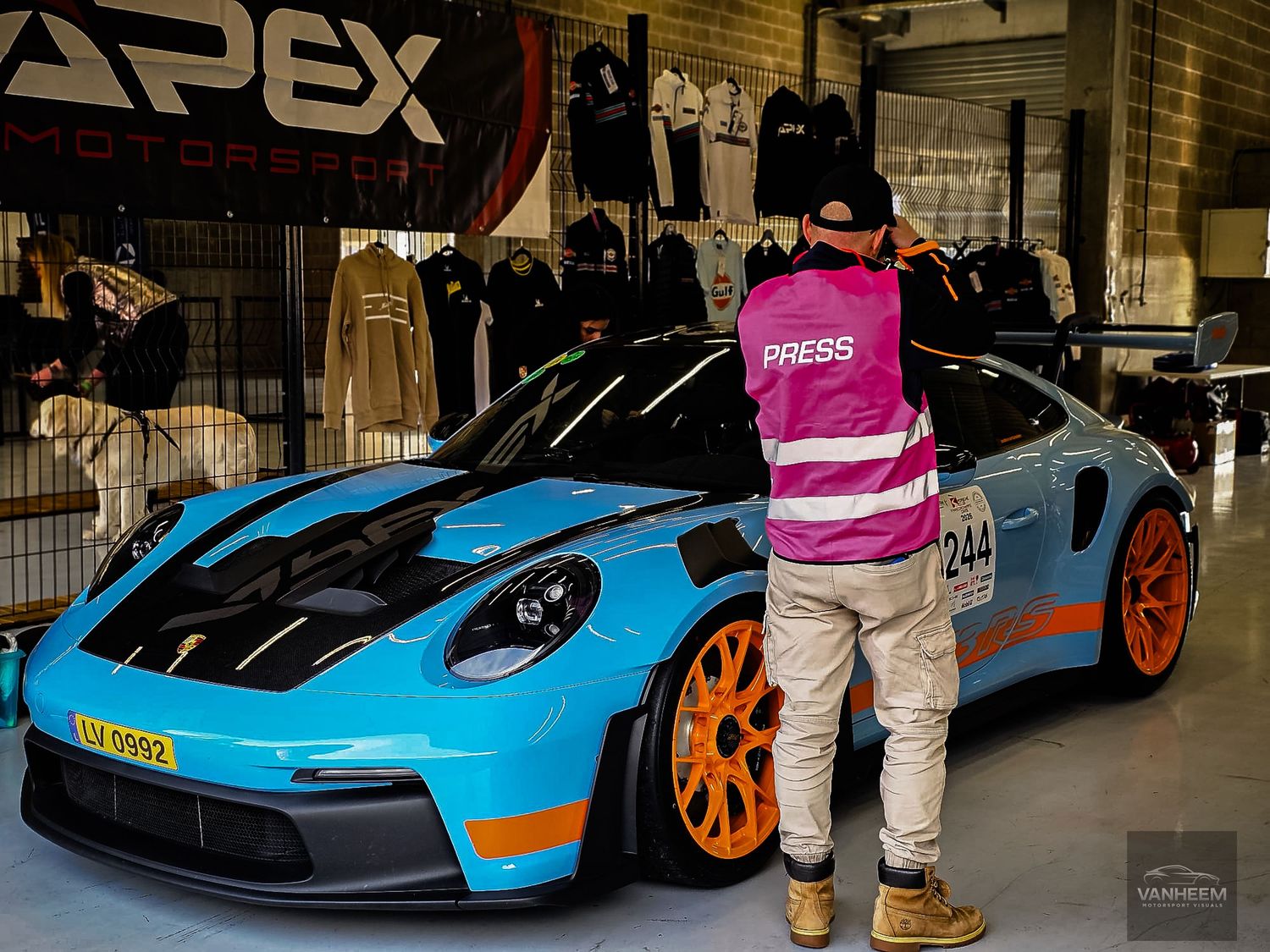 A press photographer in a pink vest stands beside a Gulf-liveried Porsche 911 GT3 RS with orange wheels at Apex Motorsport.