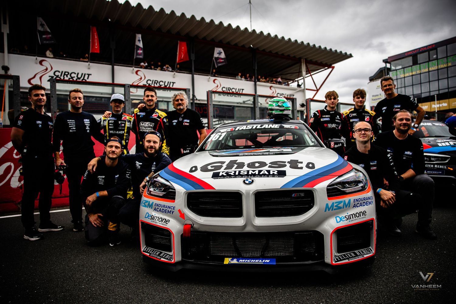 BMW M2 racing car with Ghost Kilmai livery surrounded by team members at a motorsport circuit paddock.