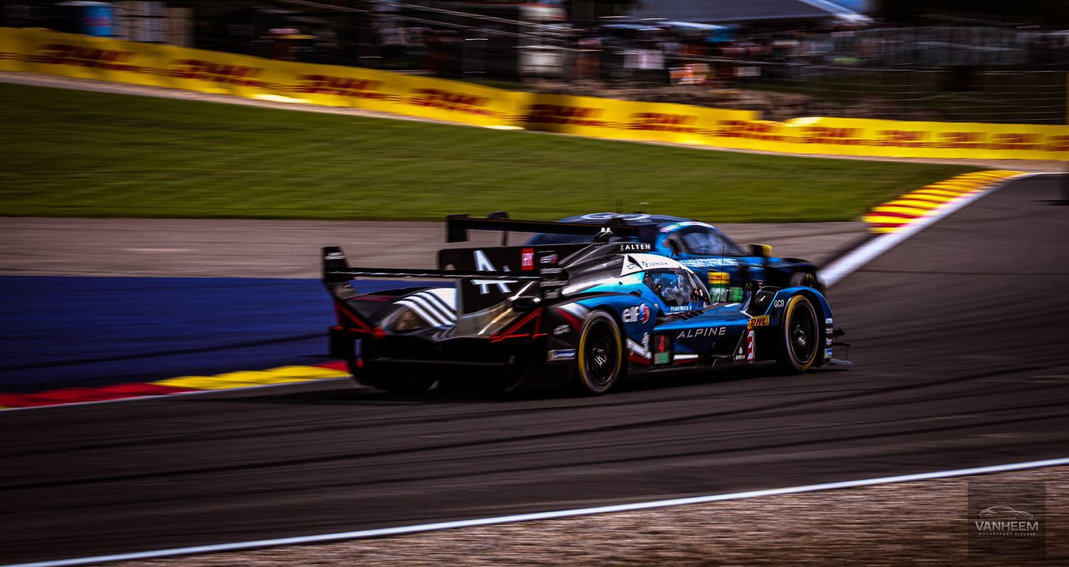 Two racing cars battle on a floodlit circuit at night, captured in dynamic motion blur during an endurance race.