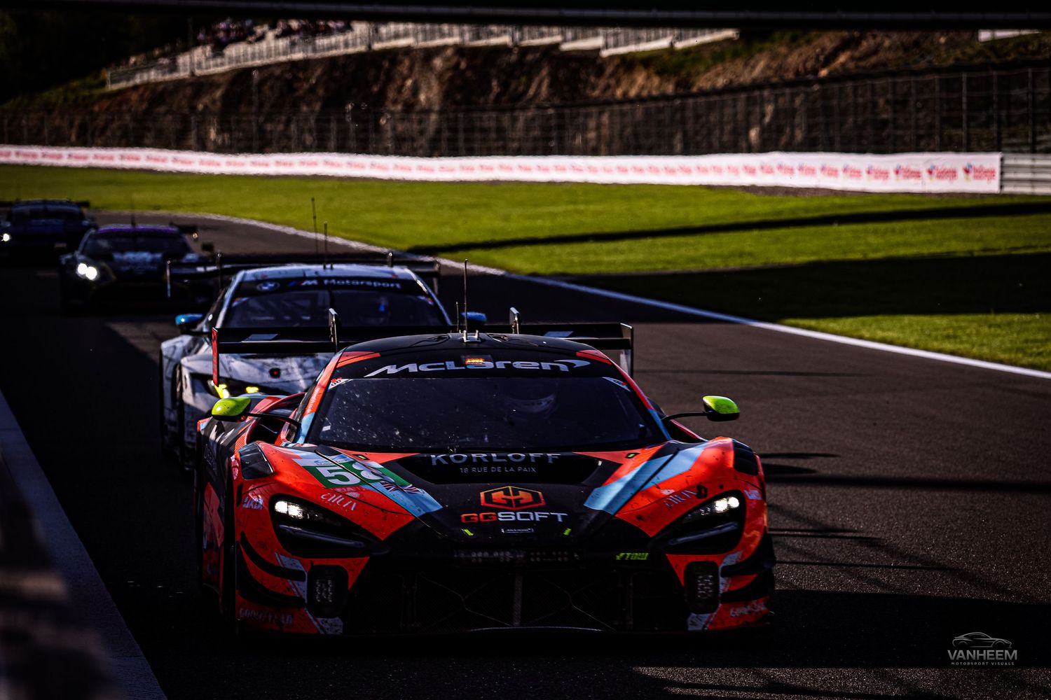 Red GT racing car leads pack on track at dusk, with McLaren visible behind during intense motorsport competition.