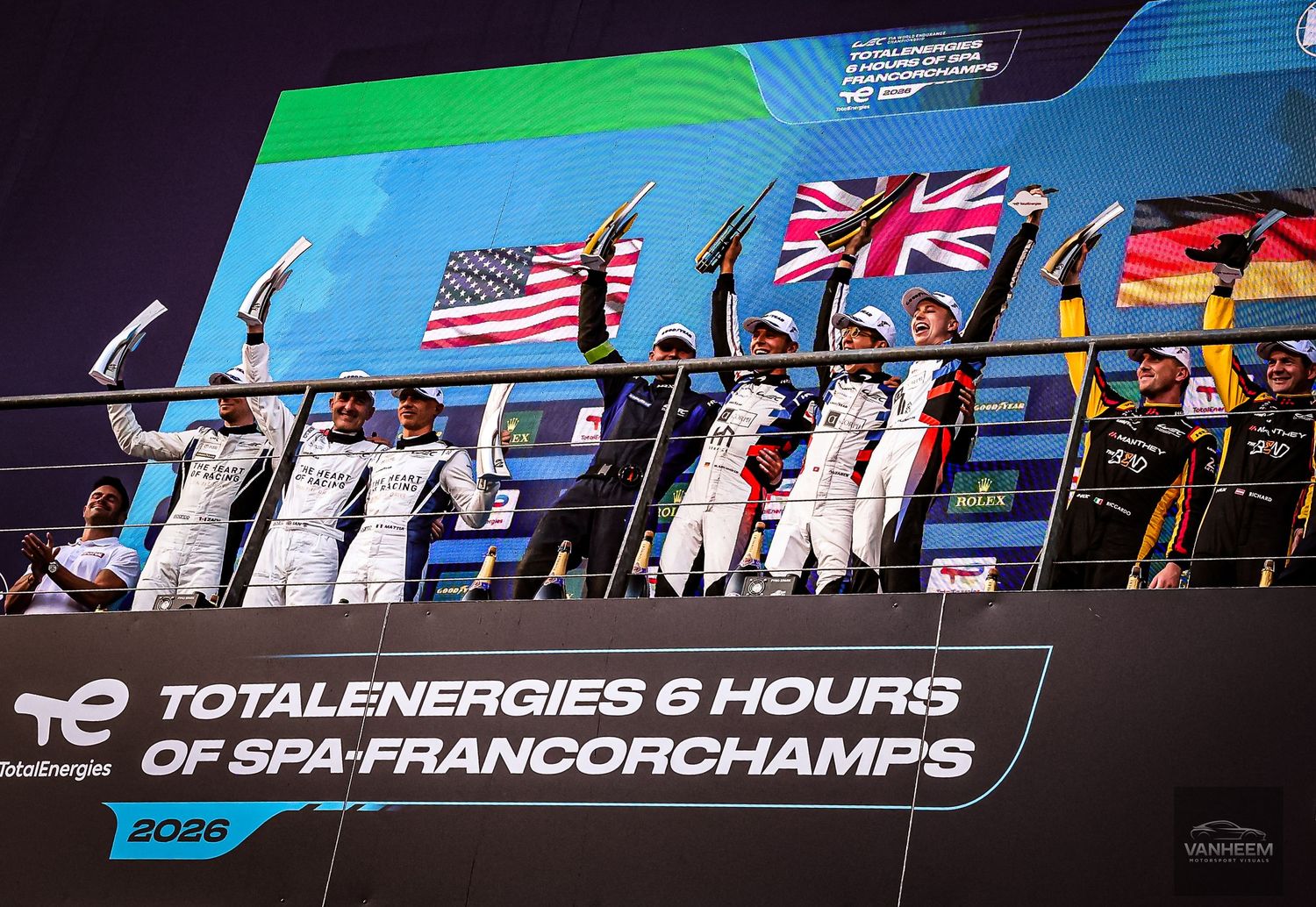 Racing teams celebrate on the podium at TotalEnergies 6 Hours of Spa-Francorchamps 2026 with USA and UK flags.