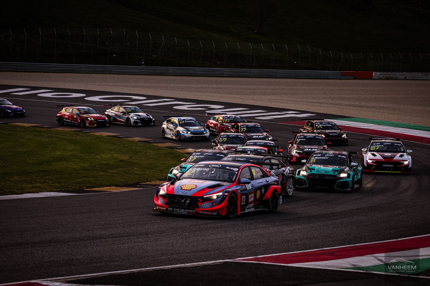 Race cars compete on a race track at dusk with colorful cars leading the pack through a turn.