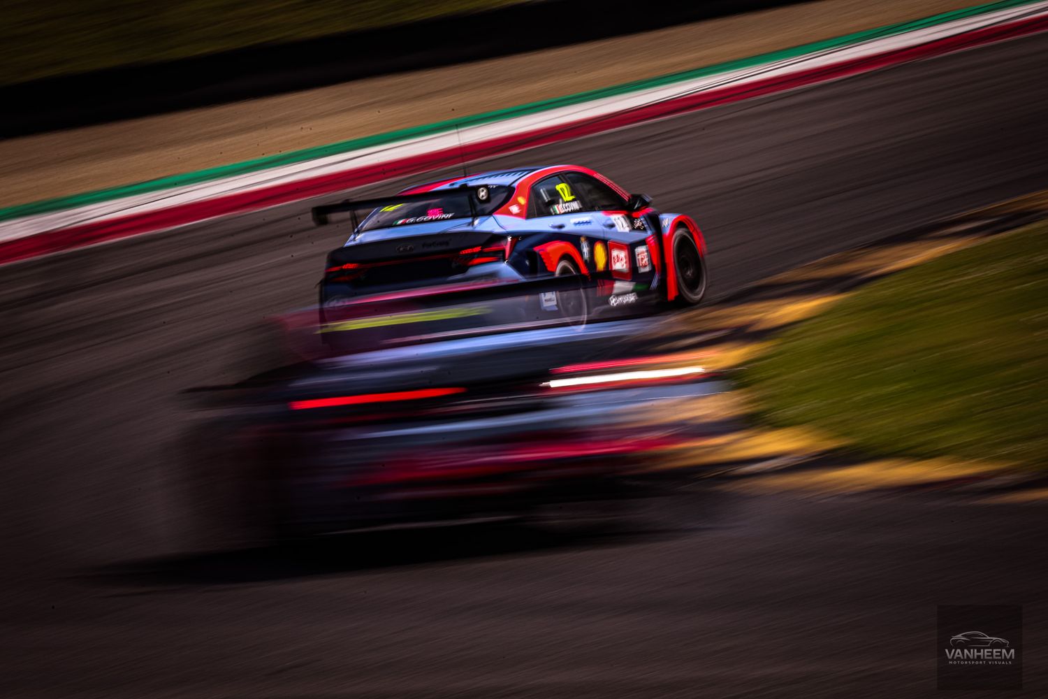 Race cars speed through a track at night with motion blur and glowing brake lights creating dramatic light trails.