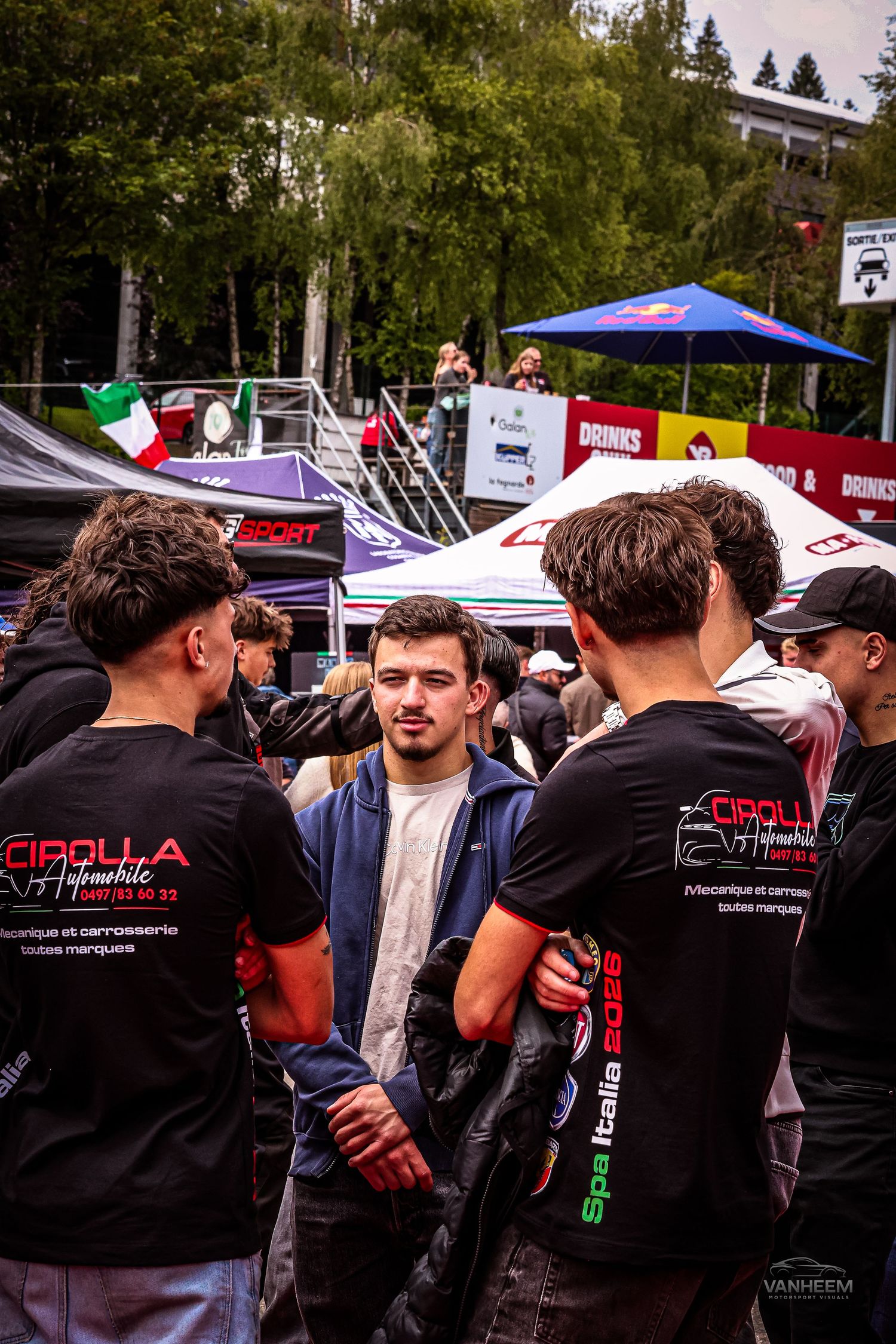 Young men in Cirolla Automobile black shirts gather at a motorsport event near Spa, Italy, with vendor tents in background.