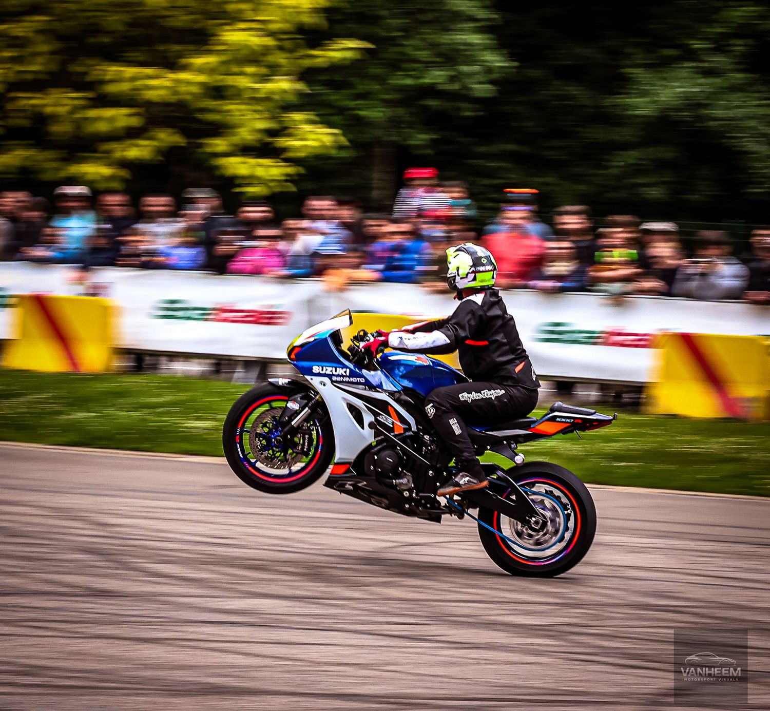 Motorcycle stunt rider performing a wheelie on a sport bike at an outdoor racing event with spectators in the background.