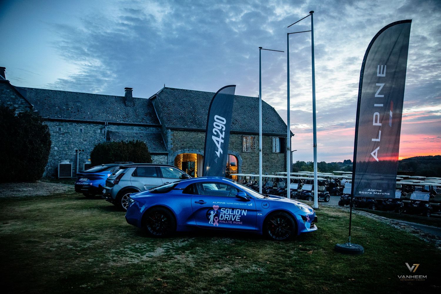Alpine A110 sports car parked at dusk near a stone building with Alpine brand flags at an automotive event.