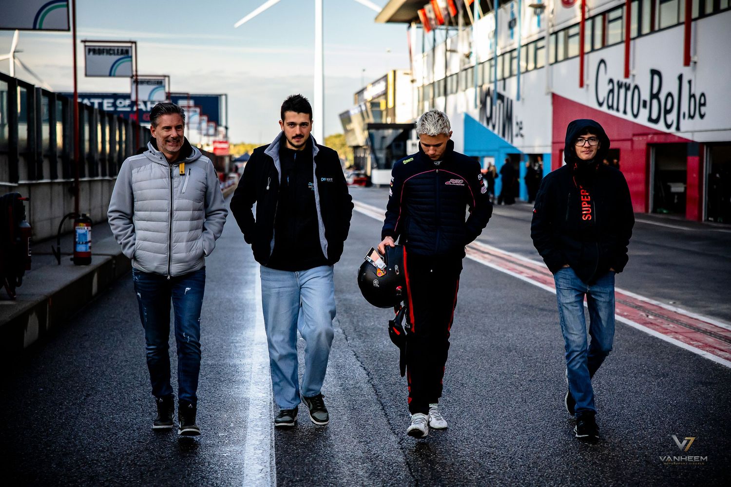 Four people walking together in a paddock area at a motorsport racing circuit on a cool day.