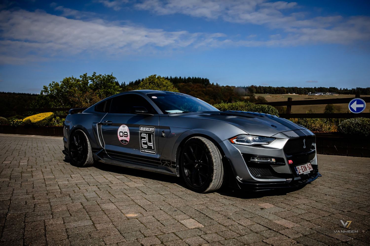 Gray Ford Mustang widebody race car with number 24 livery parked on cobblestone track under blue sky.