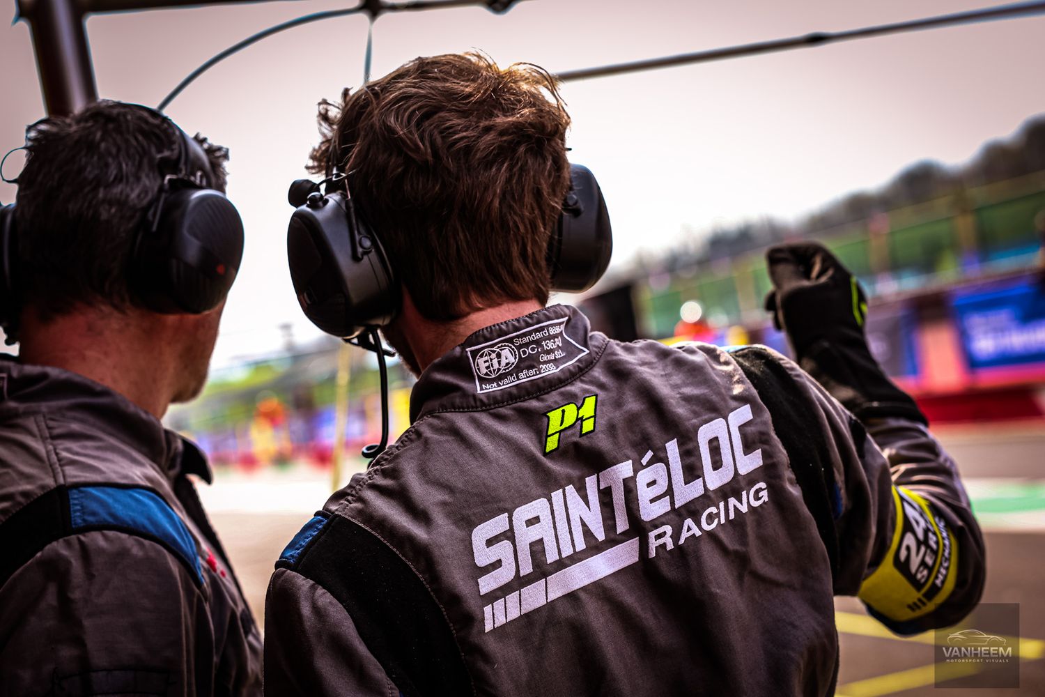 Racing team crew member wearing headphones and Saint-Eloc branded gear gestures during motorsport event.