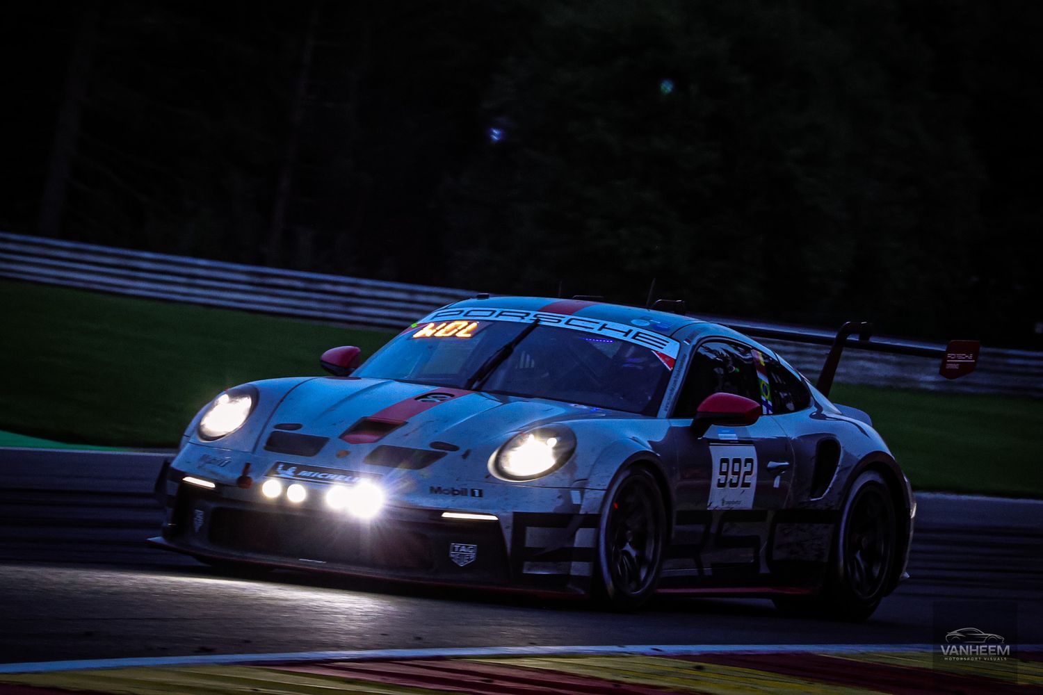 A blue Porsche 911 GT3 race car with illuminated headlights racing at night on a motorsport track.