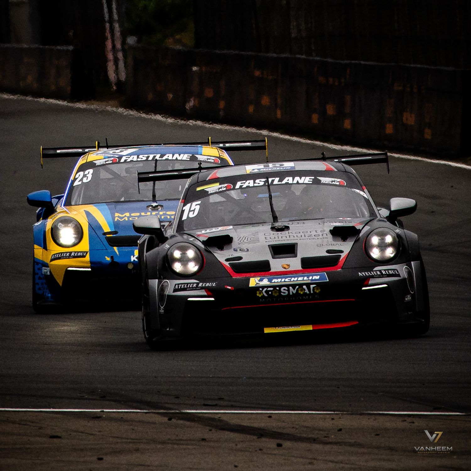 Two Porsche GT3 race cars, numbers 15 and 20, compete on track wearing Fastlane liveries during a motorsport event.