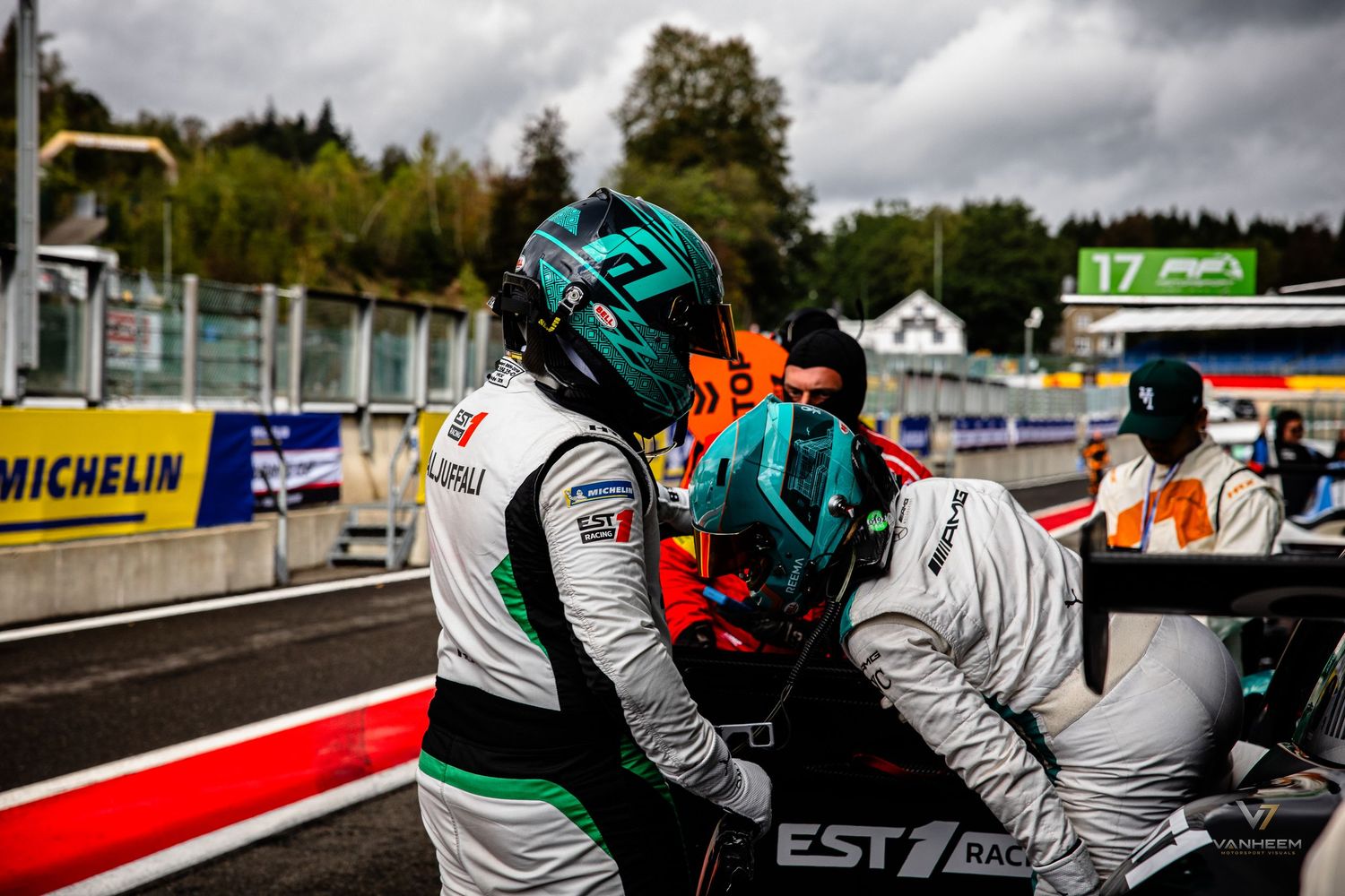 Two racing drivers in helmets and racing suits embrace on the pit lane at a motorsport circuit.