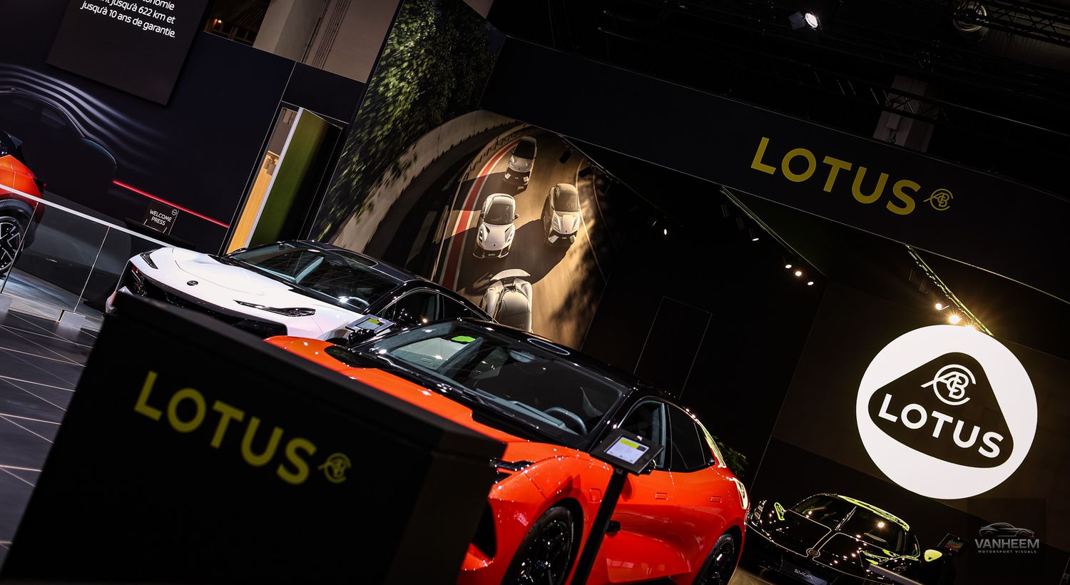 Lotus brand signage and vehicles displayed at auto show with orange accent lighting.