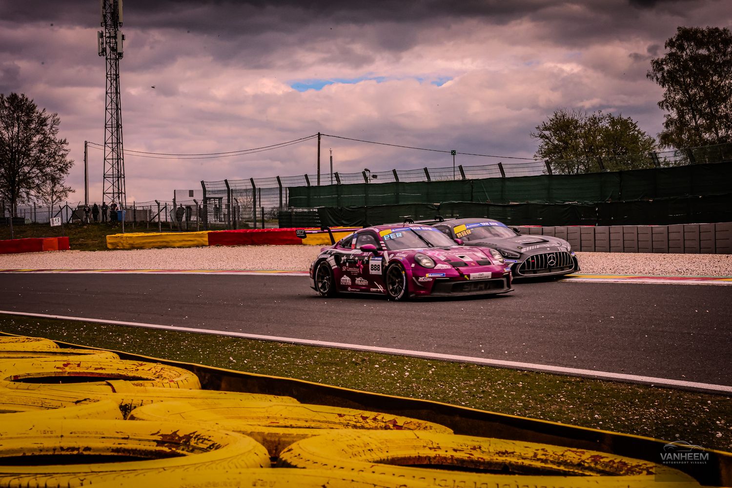 Two racing cars compete on a wet circuit track under dramatic cloudy skies during a motorsport event.