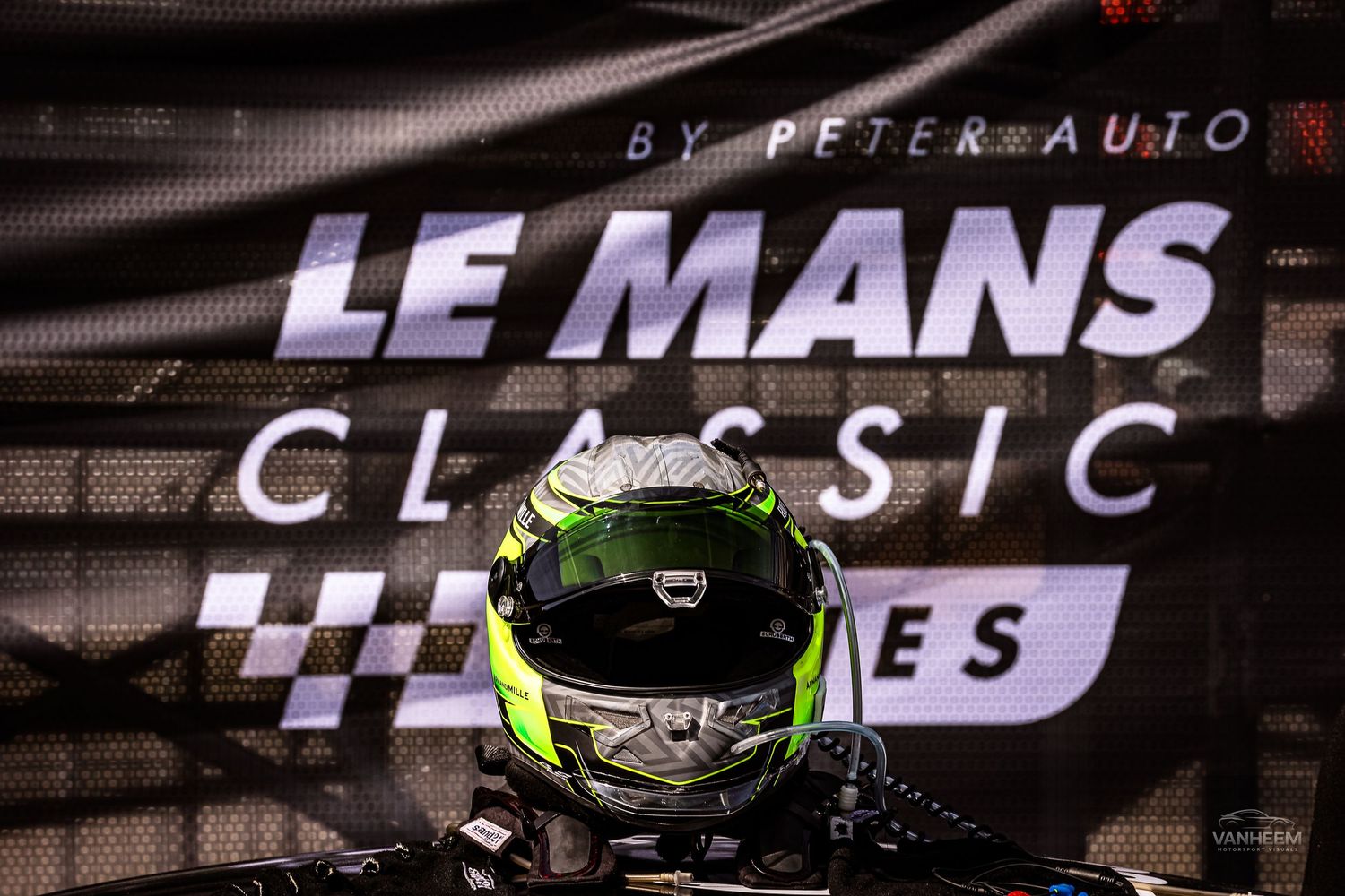 Racing driver wearing a green helmet poses in front of Le Mans Classic by Peter Auto event banner.