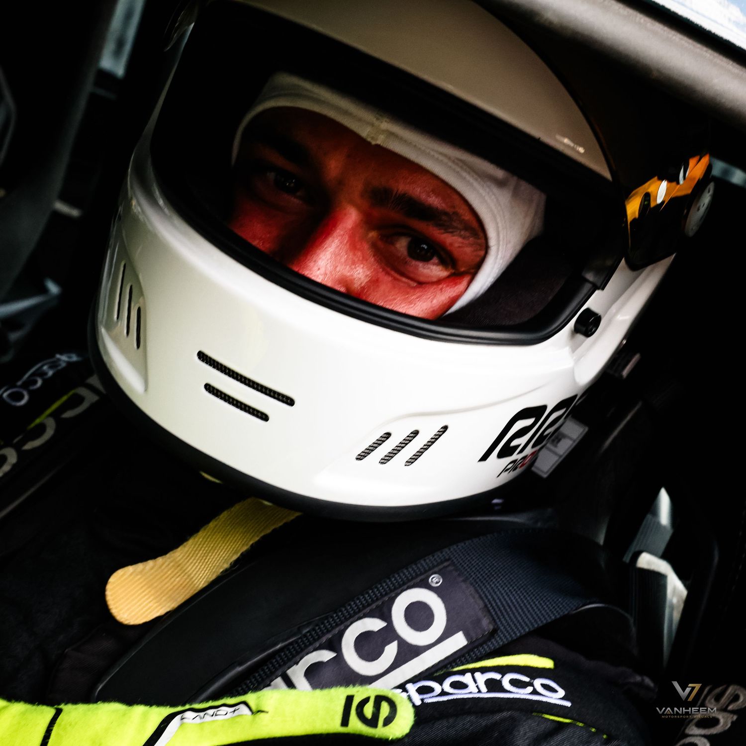 A racing driver wearing a white helmet and Sparco racing suit sits focused inside a race car cockpit.