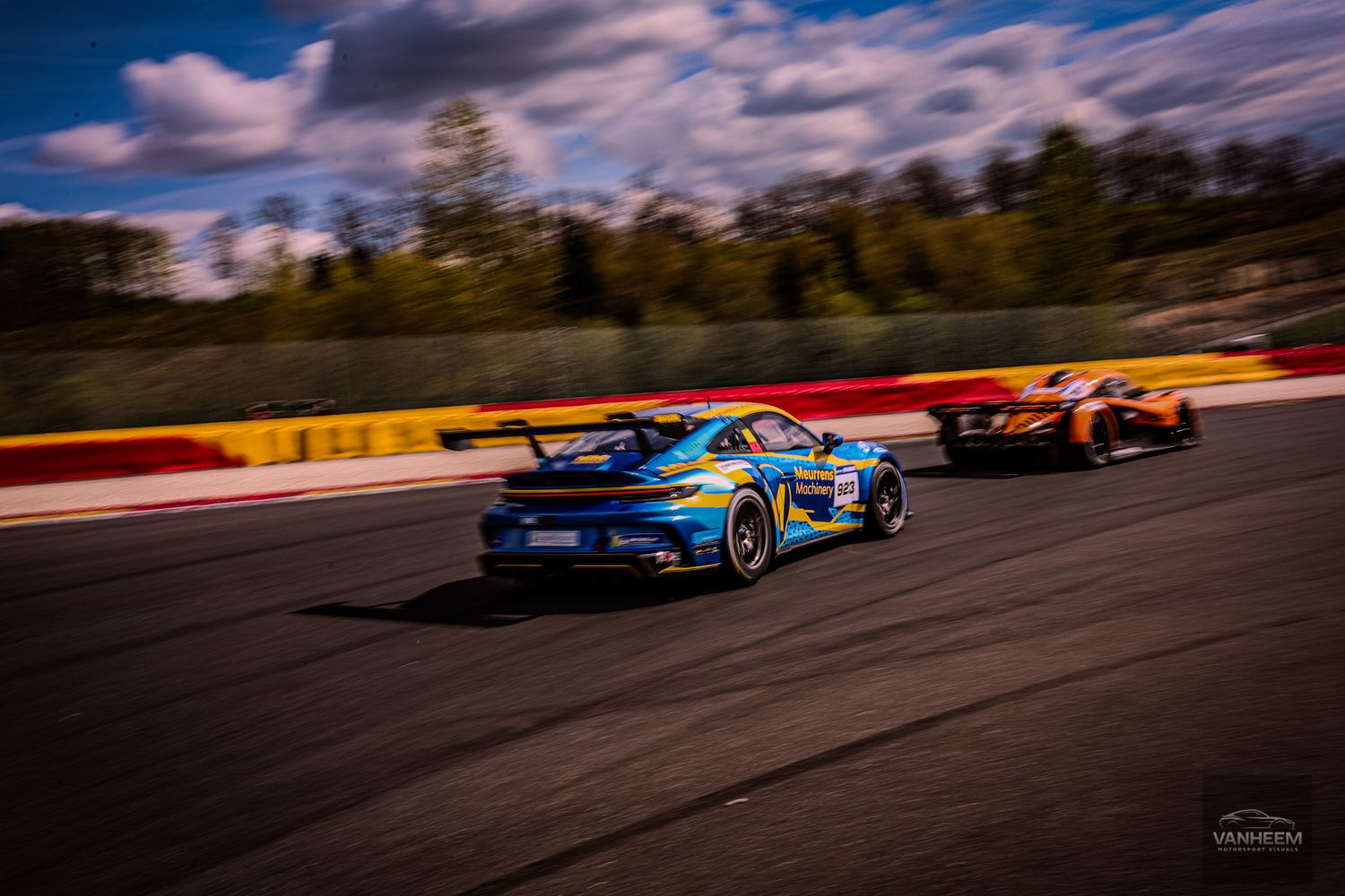 Two racing cars, a blue Porsche and orange McLaren, speeding on a track under dramatic cloudy skies.