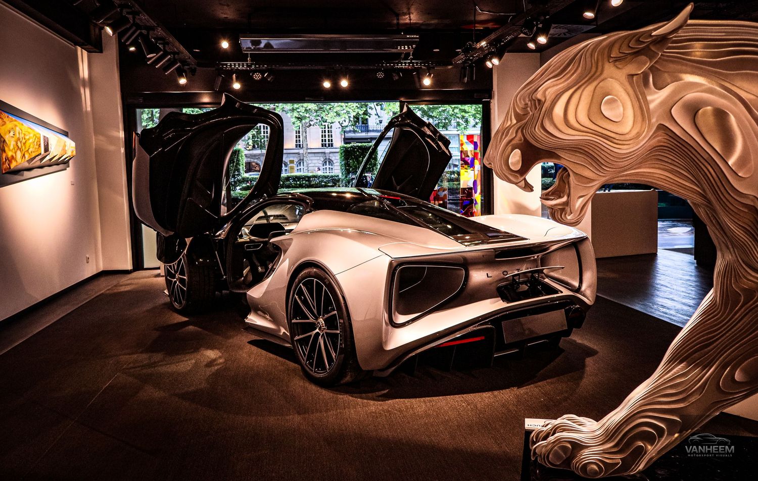 A sleek white supercar with butterfly doors open on display in a luxury gallery beside an ornate wood sculpture.