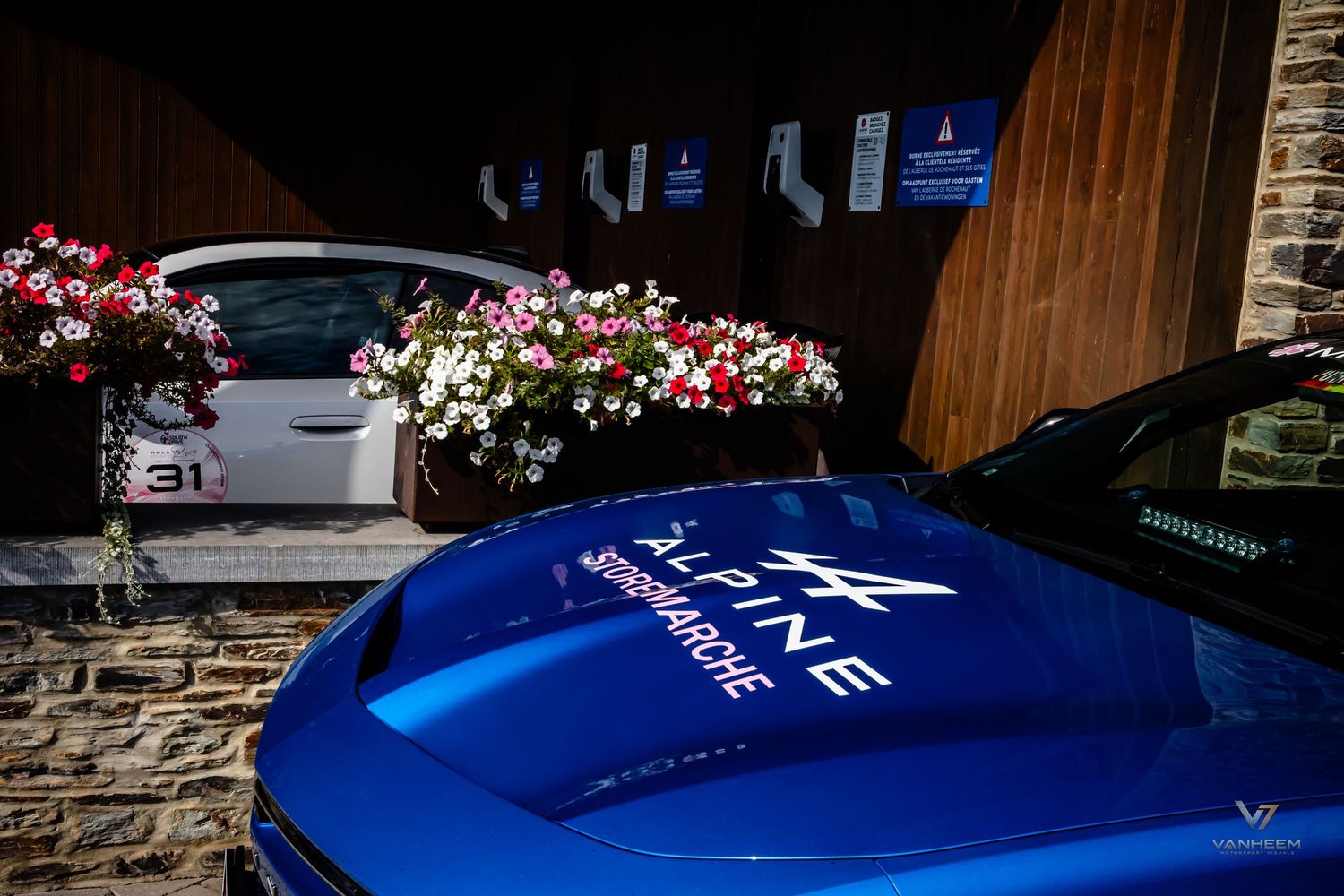 Blue Alpine A110 sports car with sponsor logos parked near a building adorned with colorful flower arrangements.