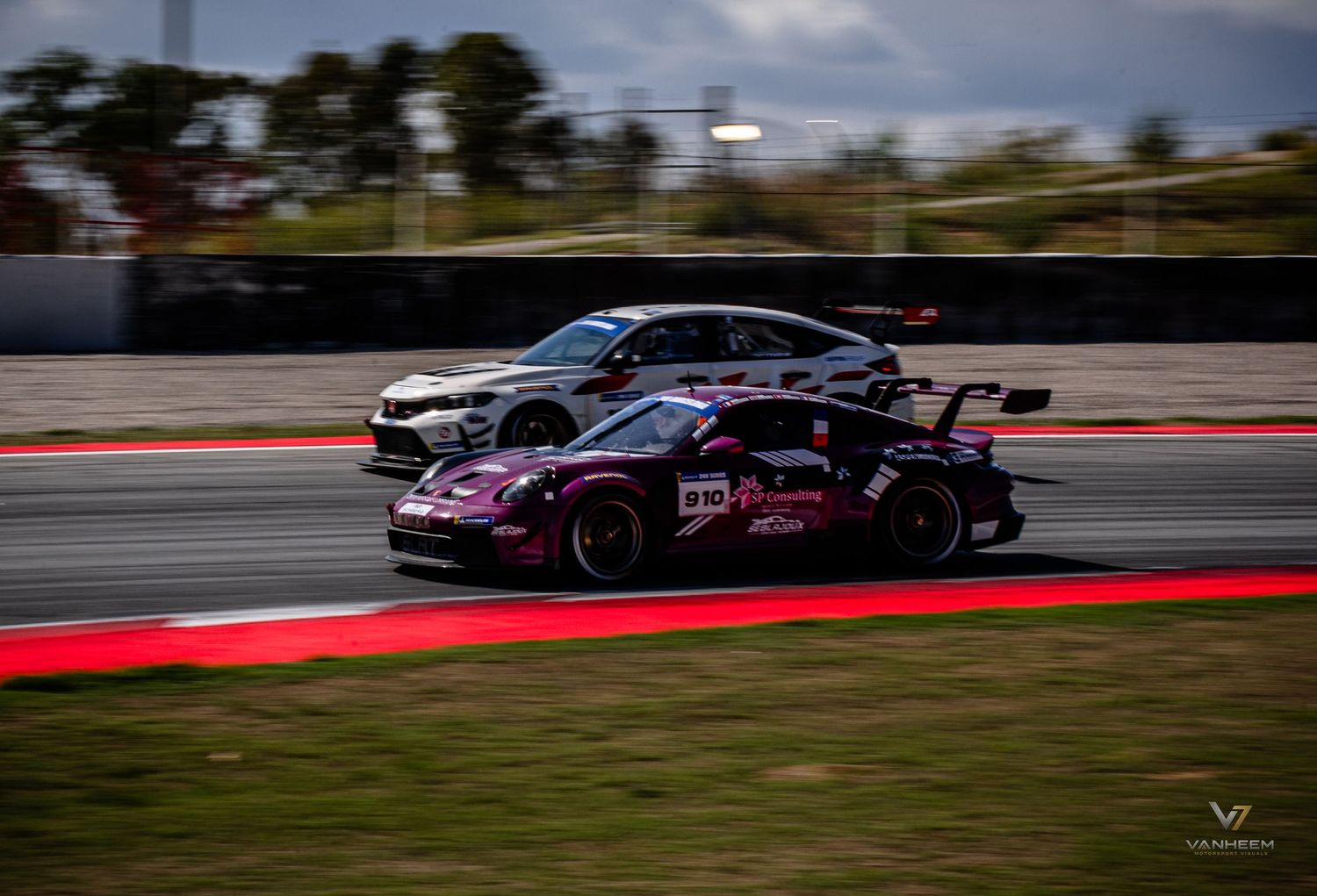 Two racing cars, a pink Porsche and a gray sedan, compete on a professional circuit track during a motorsport event.