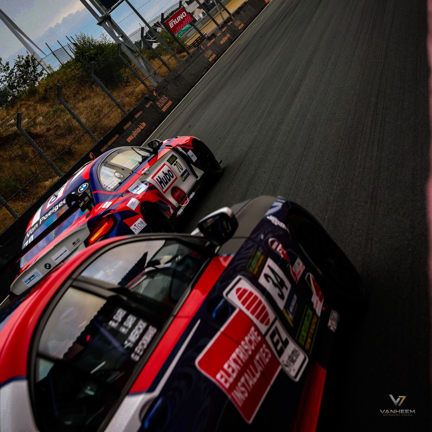 Two red and blue racing cars compete on a circuit track, photographed from a dramatic low angle perspective.