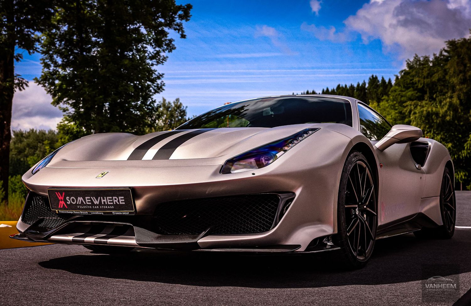 White Ferrari 488 Pista supercar with black racing stripes parked on a driveway surrounded by lush green trees.