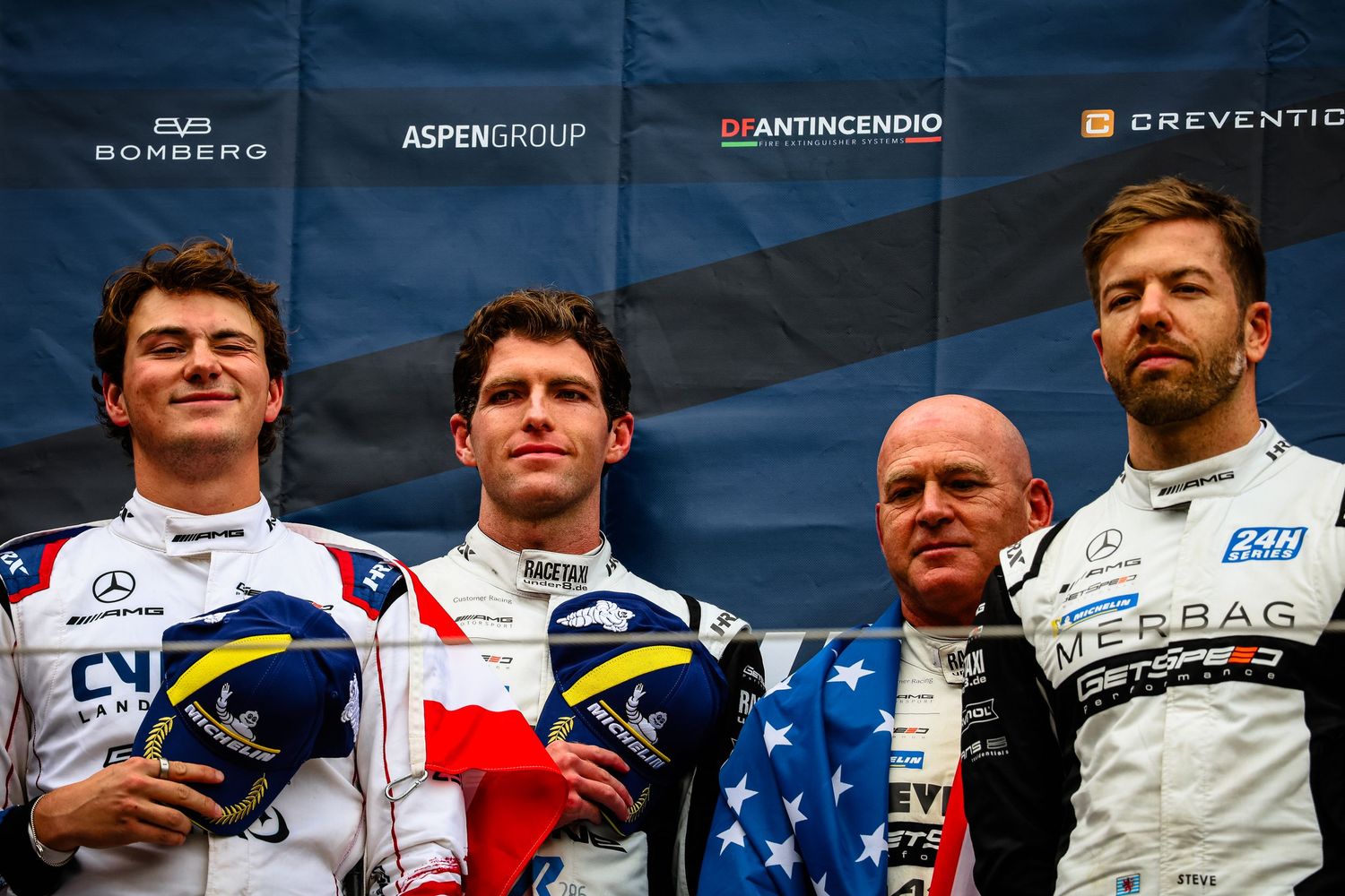 Four racing drivers pose on podium wearing team uniforms at a motorsport event with sponsor banners behind them.