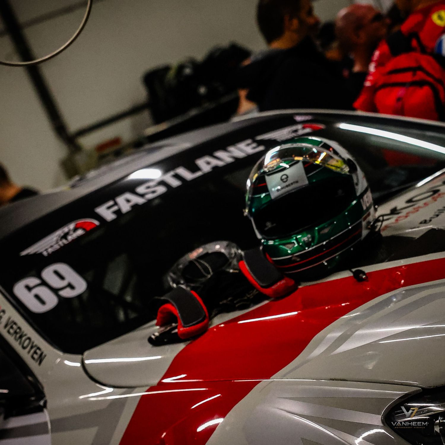 Racing car number 69 with Fastlane livery sits in pit garage with helmet resting on cockpit during motorsport event.