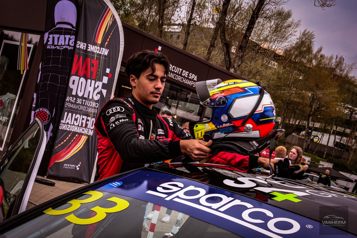 Racing driver in Audi gear adjusts helmet beside a Sparco-sponsored car numbered 33 at Circuit de Spa.