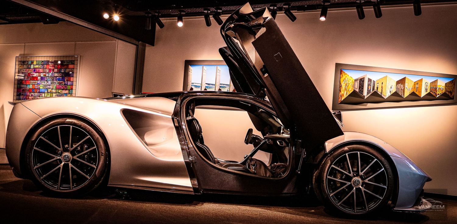 Silver supercar with scissor doors open displayed in an upscale gallery with colorful artwork on walls.