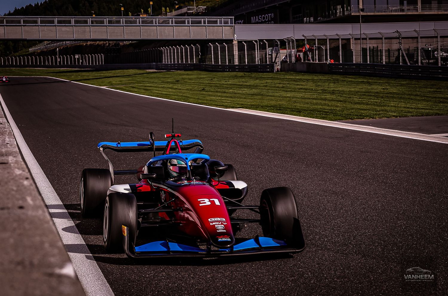 Red Formula 3 racing car number 31 speeding on a circuit track with green grass and barriers in the background.
