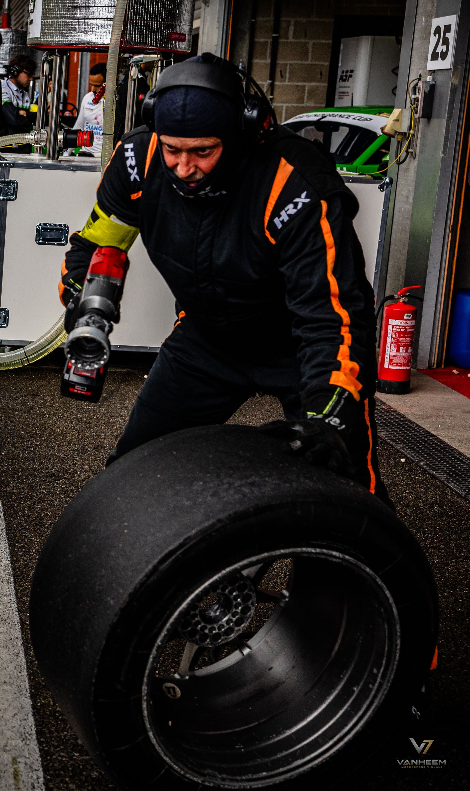 Racing mechanic in black and orange HRX suit rolling a slick tire in a pit lane garage during a motorsport event.