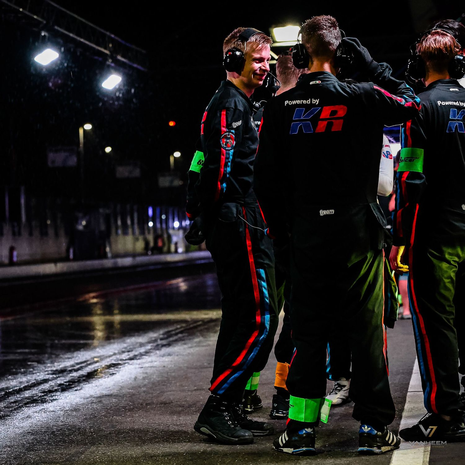 Racing team members in black and green uniforms celebrate on wet track at night under bright lights.