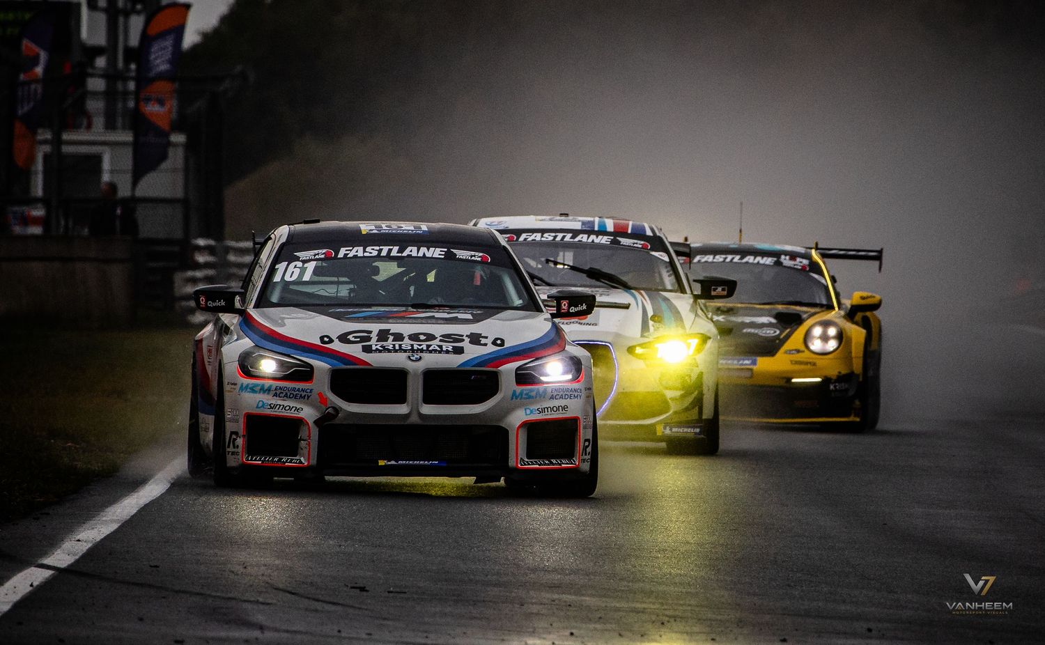 BMW M2 race car leads pack through wet track in dramatic night racing conditions at motorsport event.