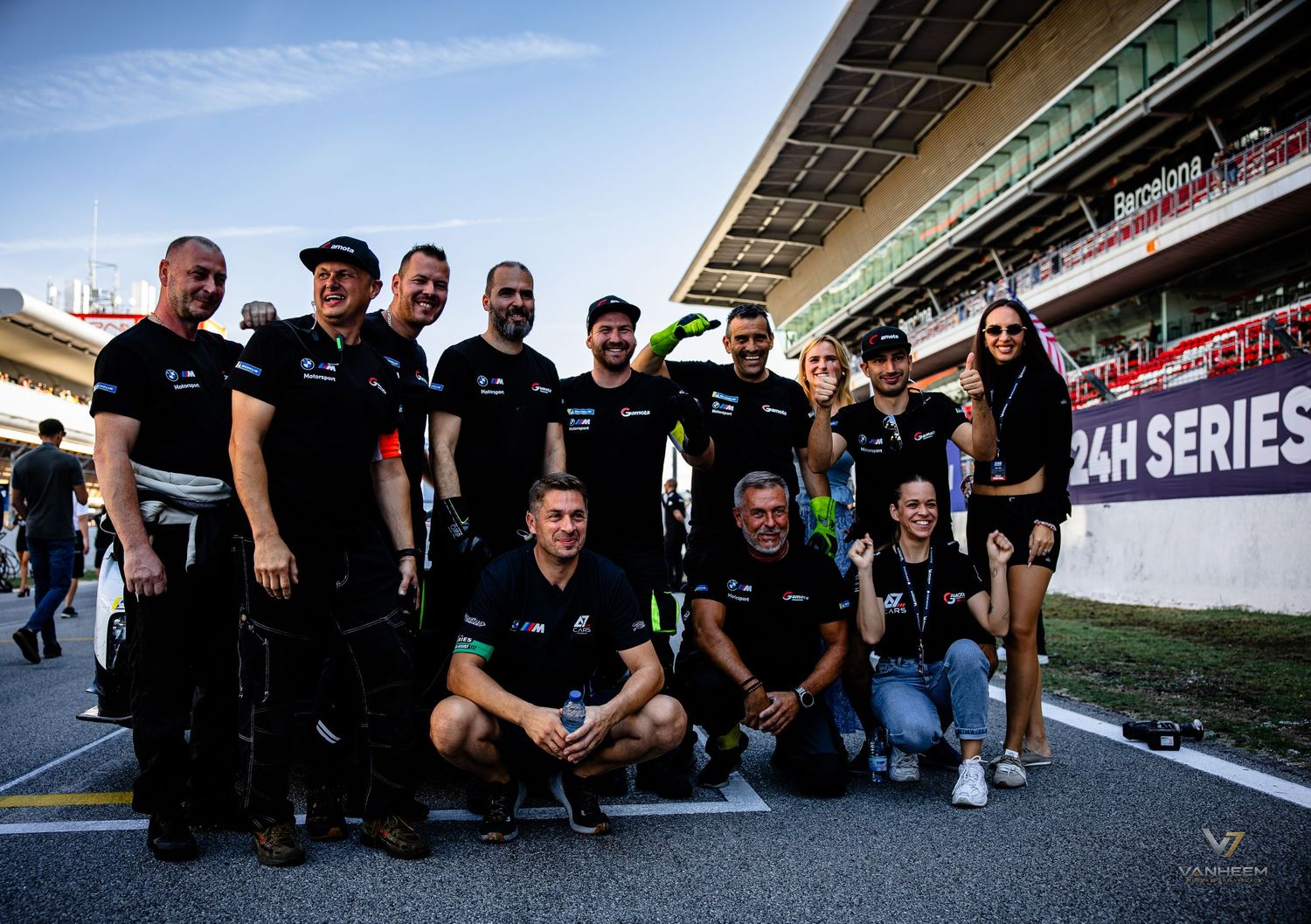 Racing team poses together at 24H Series endurance race circuit, wearing matching black uniforms on pit lane.