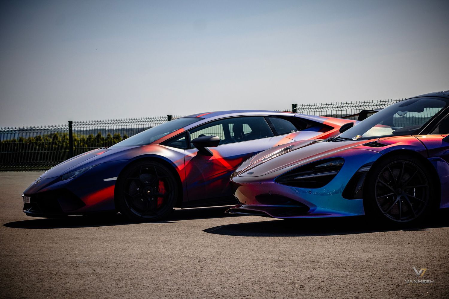 Two colorful exotic supercars with vibrant wrap designs parked on a racetrack under a clear blue sky.