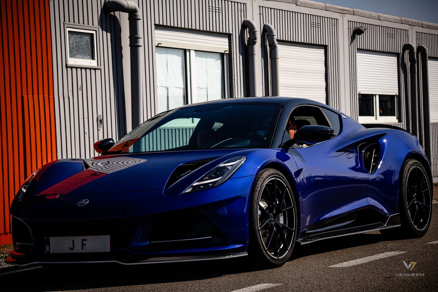 A sleek blue Lotus Emira sports car with black alloy wheels parked outside a building on a race circuit.