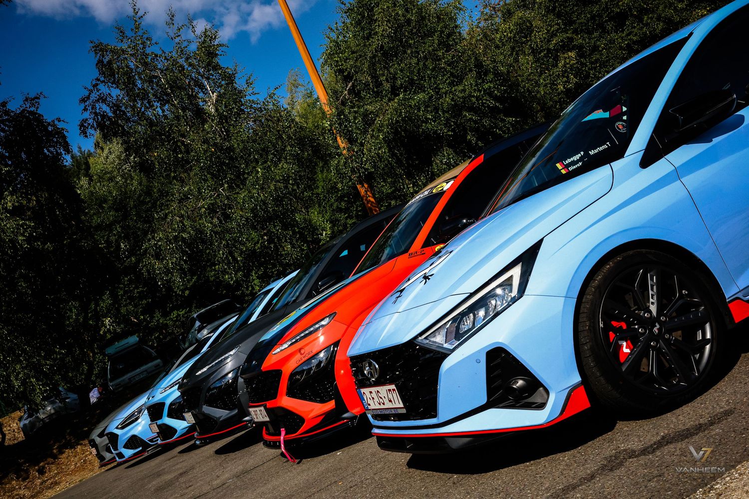 Colorful lineup of modified Hyundai i20 N sports cars parked outdoors, showcasing blue, red, and black custom paint jobs.