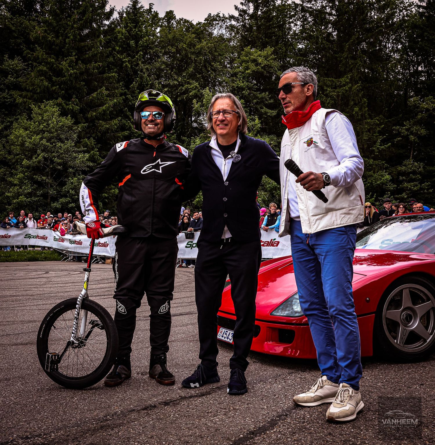 Three men posing together at a car racing event, with a red Ferrari visible in the background.