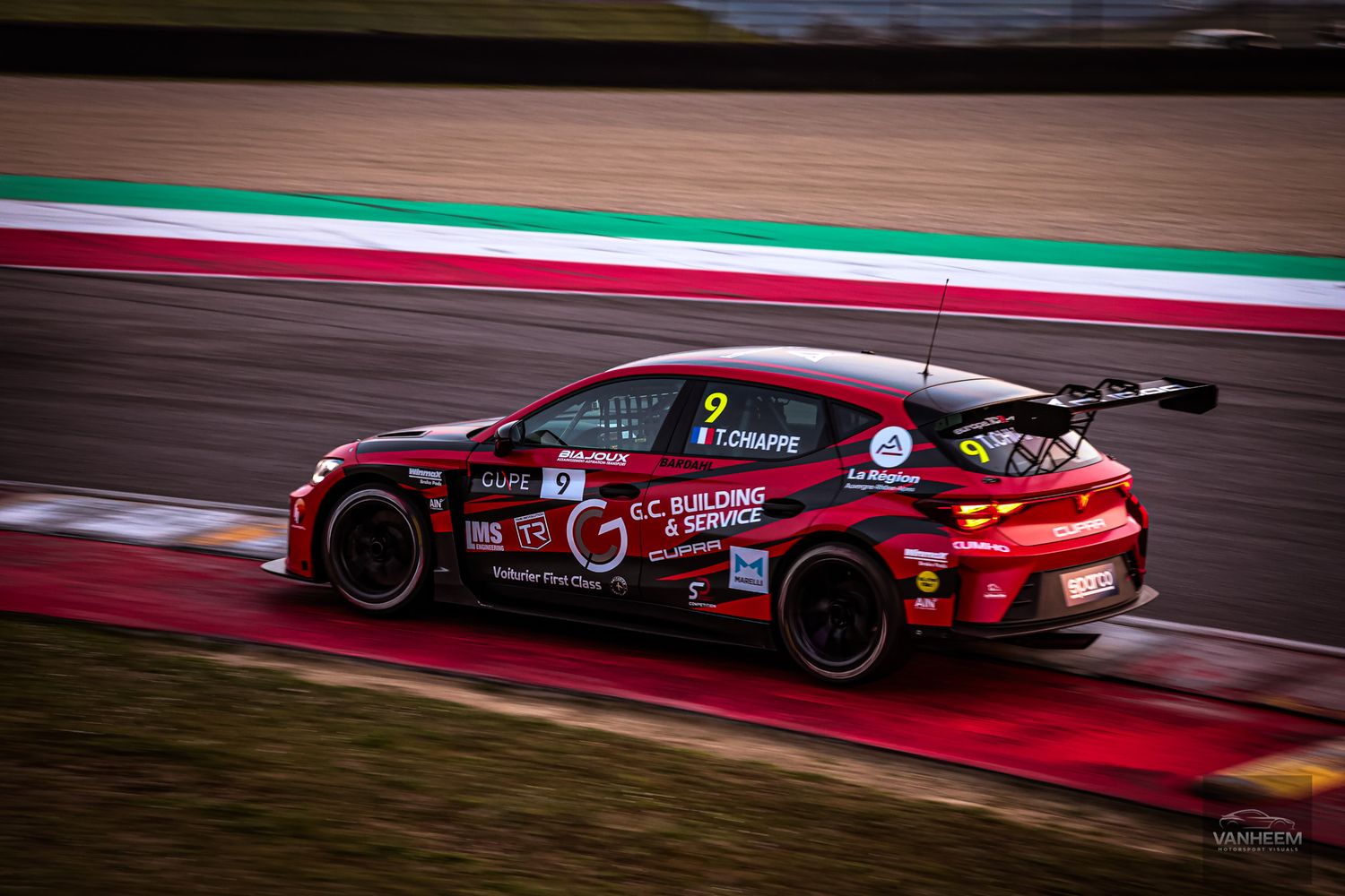 Red race car speeds around a wet track during a motorsport competition with dramatic lighting and reflections.