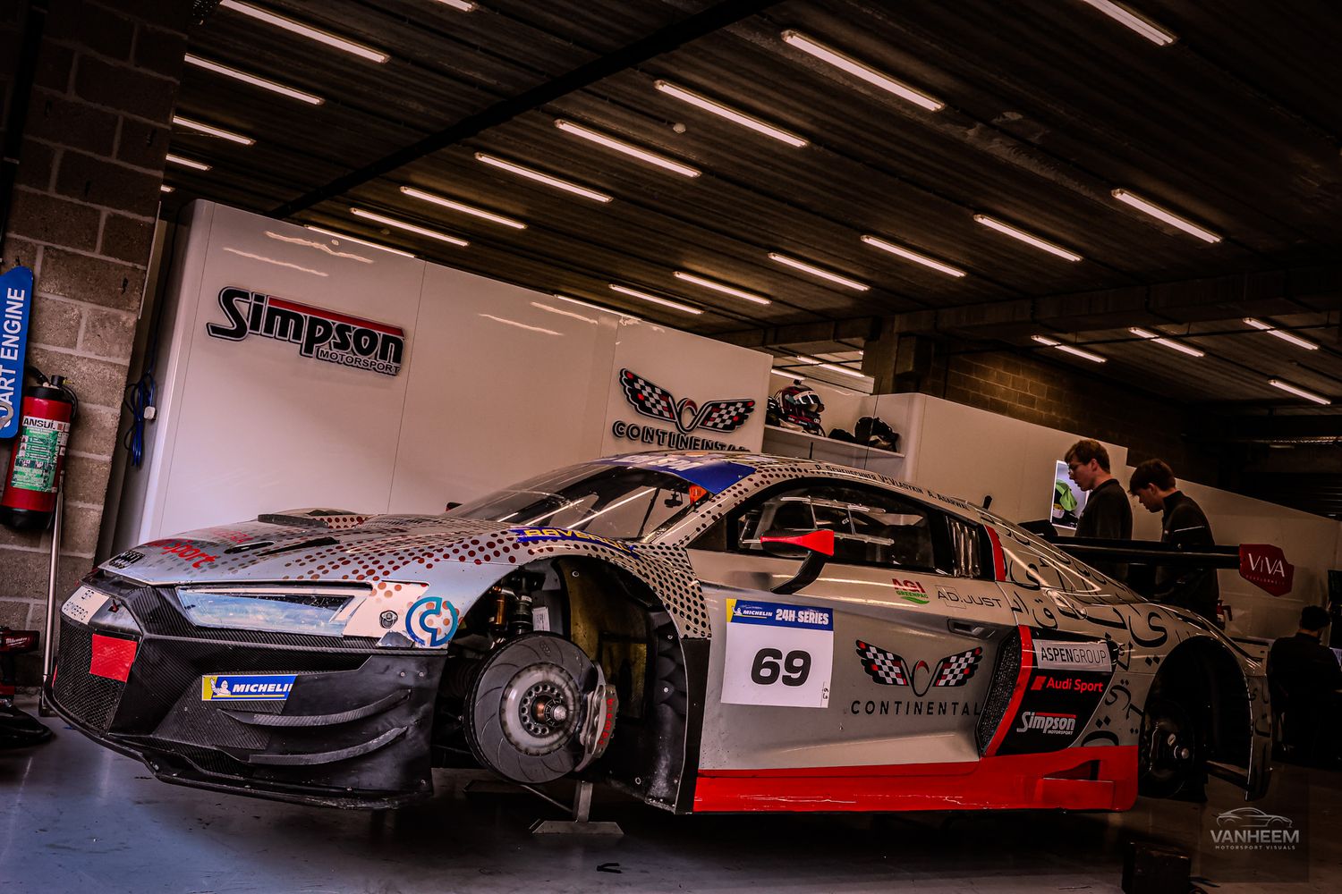 Race car #69 with camouflage livery parked in a pit garage at a motorsport event, with Simpson branding visible.