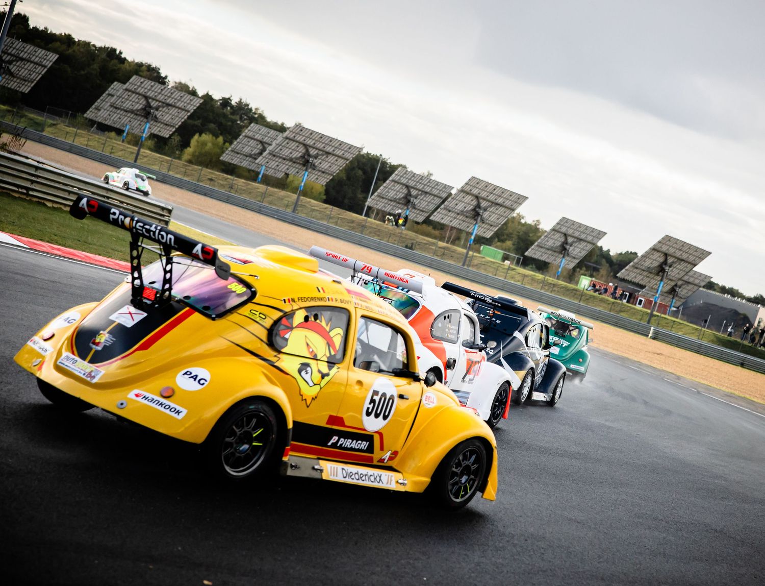 Yellow VW Beetle race car number 500 leads competitors on a racing circuit with solar panels in the background.