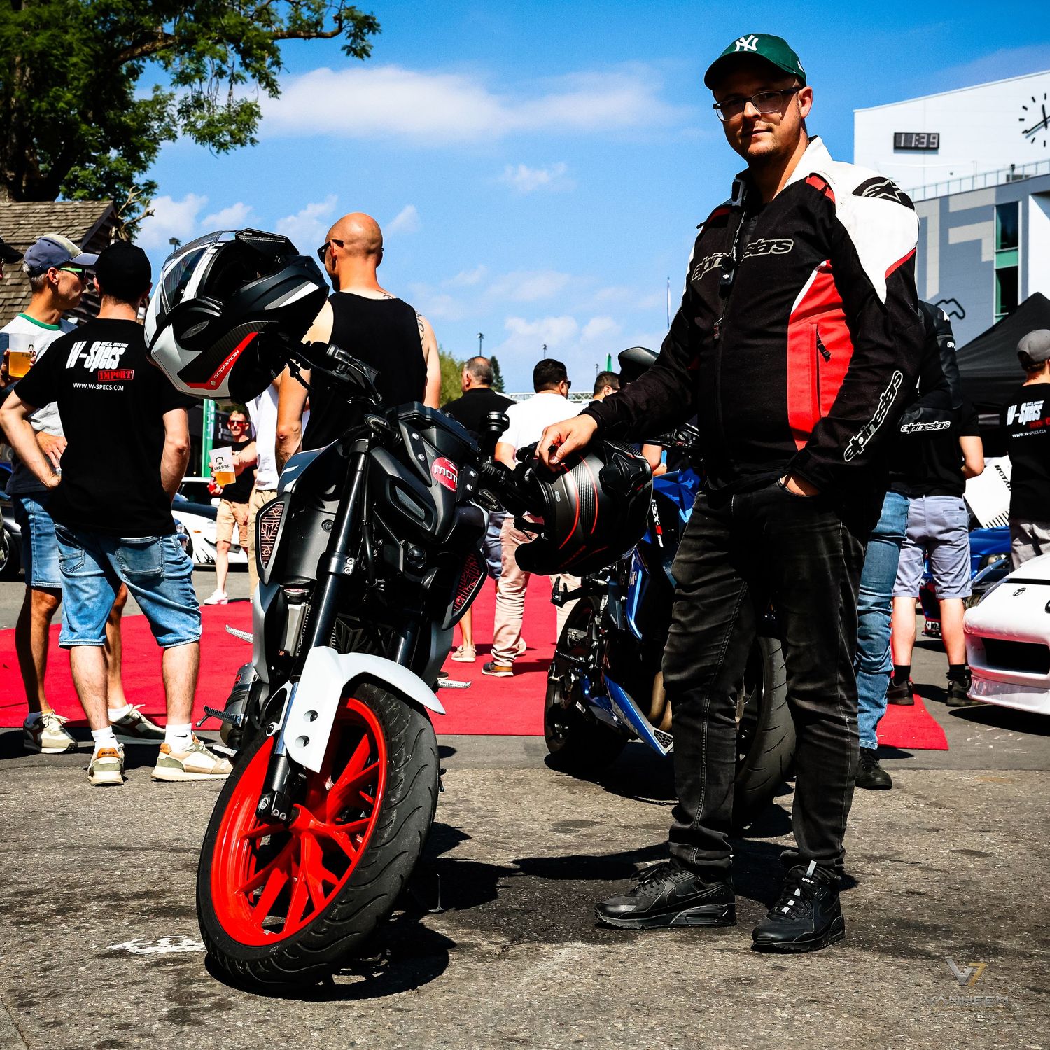A motorcyclist in black racing gear stands beside a black and red motorcycle at an outdoor motorsport event on a sunny day.