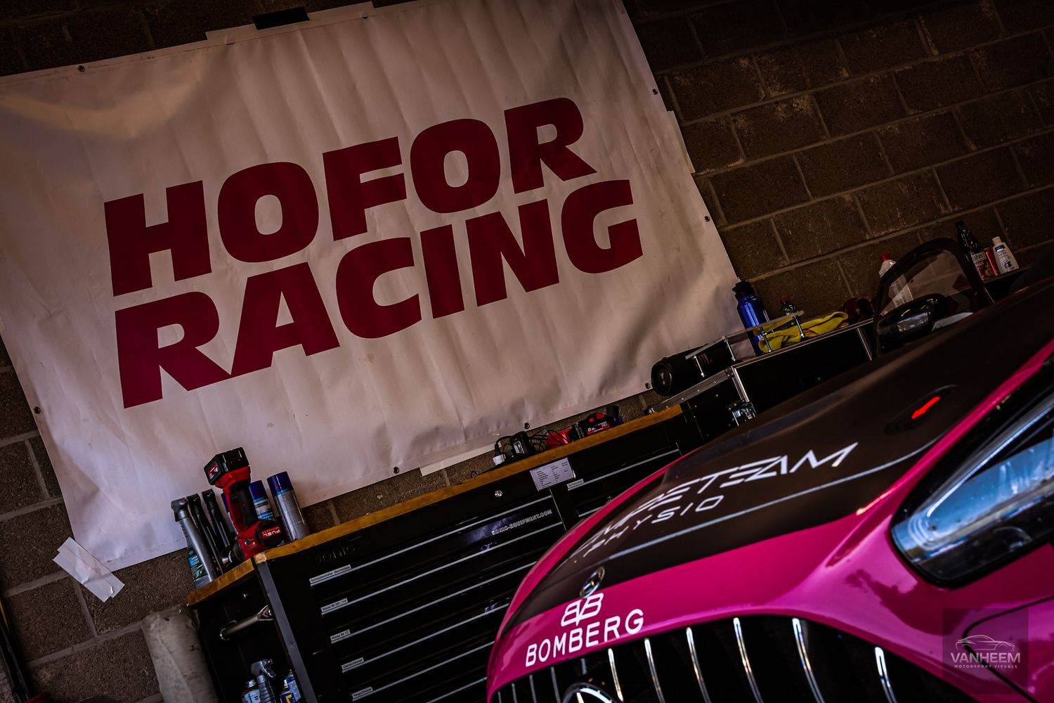 Hofor Racing team garage with a white banner, pink Mercedes-AMG GT3 race car, and tool chest visible.